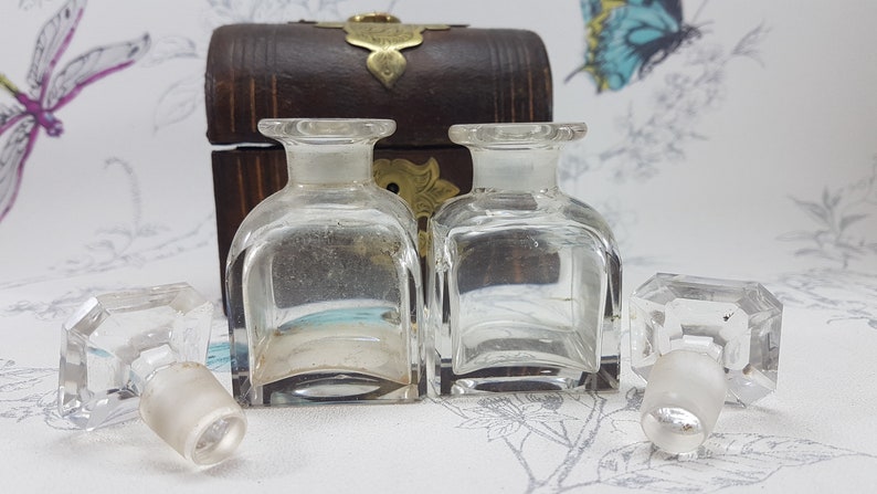 May include: Two clear glass bottles with stoppers in a wooden box with brass accents. The bottles are square shaped and have a clear liquid inside. The box is closed and has a brass latch.