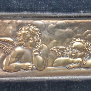 May include: A gold-colored metal plaque with two cherubs with wings, one looking to the side and the other looking straight ahead. The cherubs are set against a background of clouds.