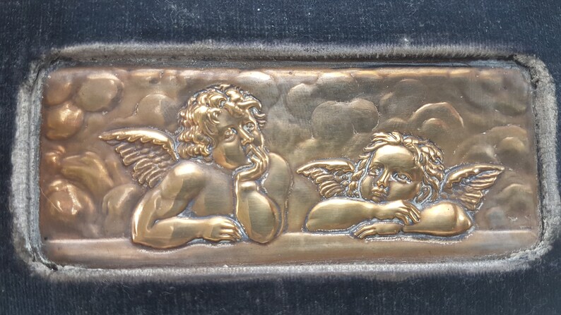 May include: A gold-colored metal plaque with two cherubs with wings, one looking to the side and the other looking straight ahead. The cherubs are set against a background of clouds.
