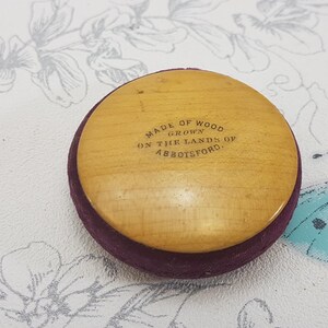May include: A round wooden button with a purple velvet backing. The button has the text "Made of Wood Grown on the Lands of Abbotsford." printed on it.