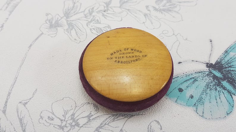 May include: A round wooden button with a purple velvet backing. The button has the text "Made of Wood Grown on the Lands of Abbotsford." printed on it.