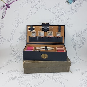 May include: A black leather sewing box with a hinged lid. The box is open and contains spools of thread, needles, and a pair of scissors. The box is sitting on a brown wooden surface.