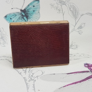 Puede incluir: Un pequeño compacto rectangular de color burdeos con un borde dorado. El compacto tiene un patrón texturizado, similar al de la piel de serpiente.