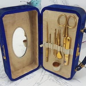 May include: A blue velvet travel case with a gold metal handle and a mirror inside. The case is open and contains a set of gold metal sewing tools, including a pair of scissors, a thimble, and needles.