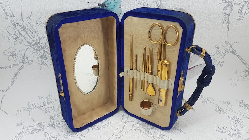May include: A blue velvet travel case with a gold metal handle and a mirror inside. The case is open and contains a set of gold metal sewing tools, including a pair of scissors, a thimble, and needles.