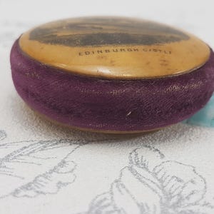 May include: A small, round, vintage pincushion with a purple velvet covering. The top is made of a cream-colored material with a black and white image of Edinburgh Castle and the text "EDINBURGH CASTLE".