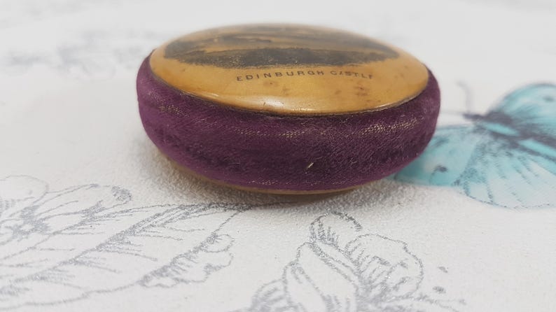 May include: A small, round, vintage pincushion with a purple velvet covering. The top is made of a cream-colored material with a black and white image of Edinburgh Castle and the text "EDINBURGH CASTLE".