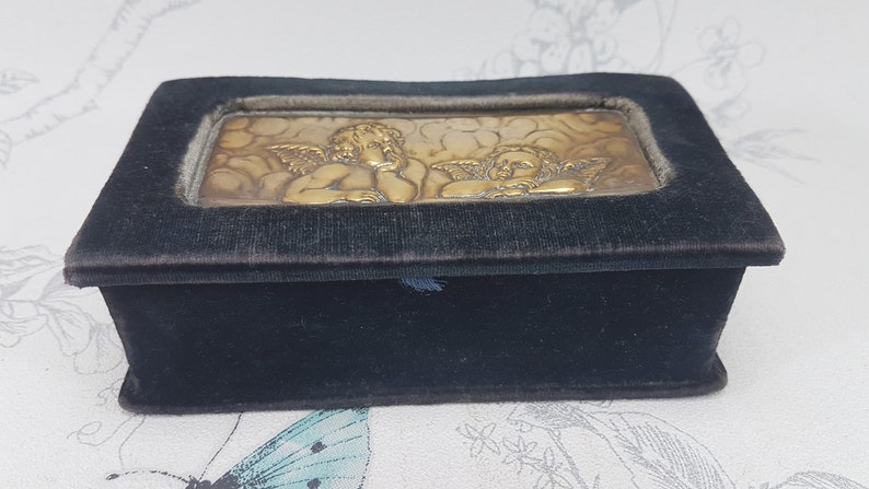 May include: A small, black velvet box with a gold-toned embossed image of two cherubs on the lid.