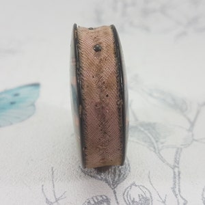 K&ouml;nnte beinhalten: Eine kleine, rechteckige, vintage M&uuml;nzgeldb&ouml;rse mit dunklem Rahmen und hellbraunem Stoffk&ouml;rper. Die B&ouml;rse hat eine leicht gebogene Form und einen kleinen Metallverschluss. Die Au&szlig;enseite zeigt ein verblasstes Blumenmuster.