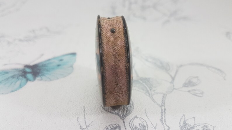 K&ouml;nnte beinhalten: Eine kleine, rechteckige, vintage M&uuml;nzgeldb&ouml;rse mit dunklem Rahmen und hellbraunem Stoffk&ouml;rper. Die B&ouml;rse hat eine leicht gebogene Form und einen kleinen Metallverschluss. Die Au&szlig;enseite zeigt ein verblasstes Blumenmuster.