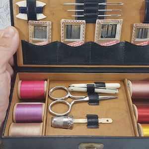 May include: A black leather sewing kit with a variety of sewing supplies, including spools of thread in pink, purple, beige, red, and yellow, a pair of scissors, a thimble, and needles in small packages.