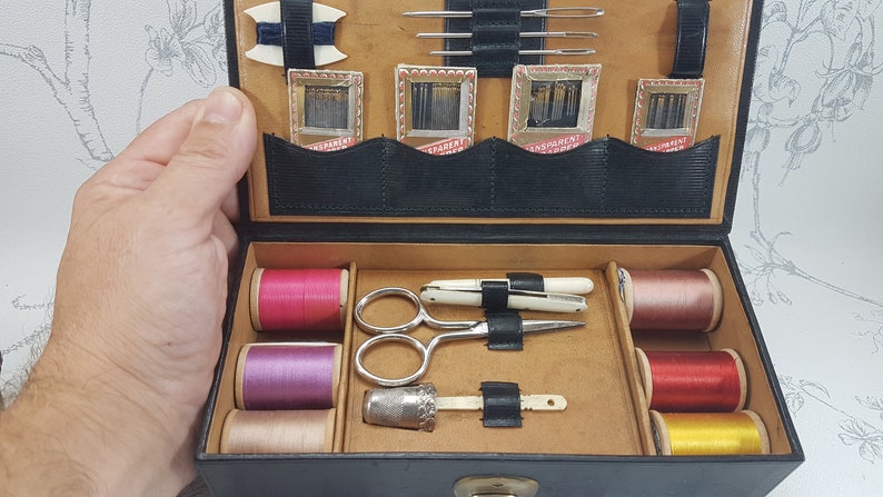 May include: A black leather sewing kit with a variety of sewing supplies, including spools of thread in pink, purple, beige, red, and yellow, a pair of scissors, a thimble, and needles in small packages.