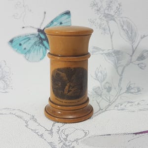 Puede incluir: Un recipiente de madera de color marrón claro, con un cuerpo cilíndrico, una parte superior y una base redondeadas. El recipiente tiene una imagen oscura en la parte delantera con texto debajo.