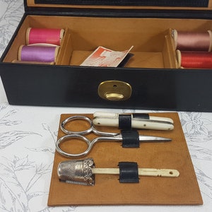 May include: A black leather sewing box with a brass latch. The box is open and contains spools of thread in pink, purple, and red. The box also contains a small pair of scissors, a needle threader, and a thimble.