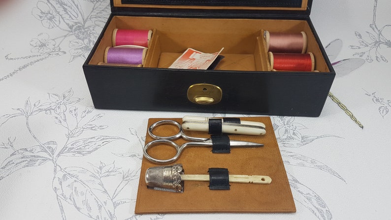 May include: A black leather sewing box with a brass latch. The box is open and contains spools of thread in pink, purple, and red. The box also contains a small pair of scissors, a needle threader, and a thimble.