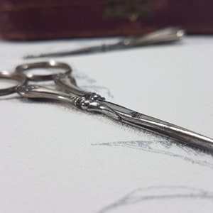 May include: A pair of silver-toned, ornate scissors with a decorative handle. The scissors are closed and lying on a white surface.