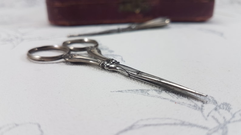 May include: A pair of silver-toned, ornate scissors with a decorative handle. The scissors are closed and lying on a white surface.