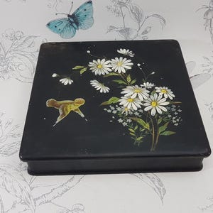 Könnte beinhalten: Eine schwarze rechteckige Schachtel mit einem gemalten Blumendesign auf der Oberseite. Das Design zeigt weiße Gänseblümchen und einen gelben Vogel auf schwarzem Hintergrund.