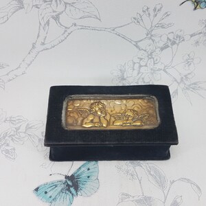 May include: A small black velvet box with a gold cherub design on the front. The box is closed and has a hinged lid.