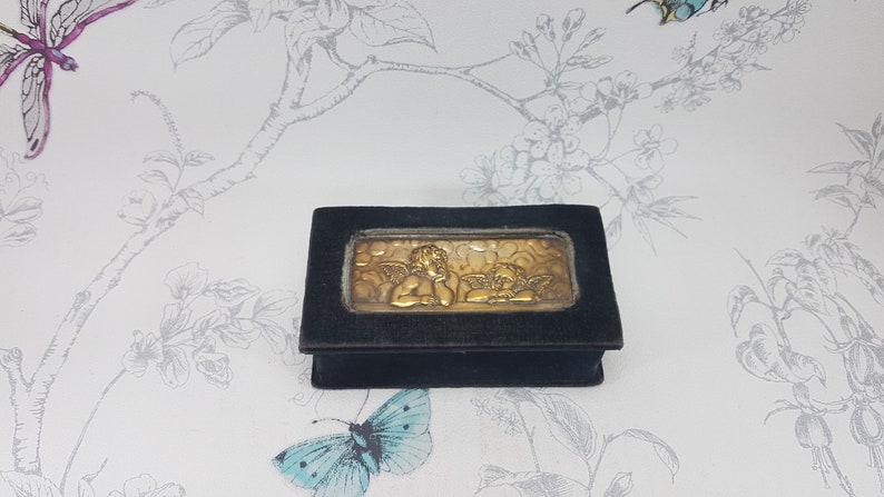 May include: A small black velvet box with a gold cherub design on the front. The box is closed and has a hinged lid.