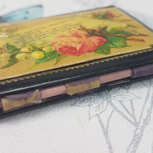 May include: A vintage black leather-bound notebook with a floral design on the cover. The cover features a pink rose, green leaves, and white flowers. The notebook is closed and has a purple ribbon tied around it.