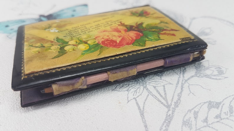 May include: A vintage black leather-bound notebook with a floral design on the cover. The cover features a pink rose, green leaves, and white flowers. The notebook is closed and has a purple ribbon tied around it.