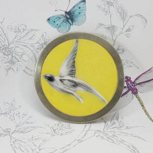 Puede incluir: Un broche redondo plateado con un fondo amarillo y un pájaro blanco en vuelo. El pájaro está pintado en un estilo realista con plumas grises y blancas.