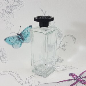 Può includere: Una bottiglia di vetro trasparente con un tappo nero. La bottiglia ha una forma quadrata con una superficie testurizzata. La bottiglia è su uno sfondo bianco con un motivo floreale.