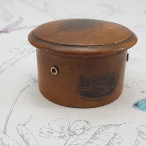 Puede incluir: Una pequeña caja redonda de madera marrón con tapa. La caja tiene un cuerpo cilíndrico y una tapa ligeramente abovedada. Una pequeña imagen está impresa en el lateral. La caja es probablemente vintage o antigua.
