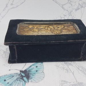 May include: A small, rectangular velvet box with a gold-toned metal inlay depicting a floral design. The box is dark blue and has a smooth, velvety texture.