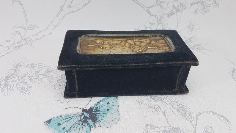 May include: A small, rectangular velvet box with a gold-toned metal inlay depicting a floral design. The box is dark blue and has a smooth, velvety texture.