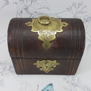 May include: A small, brown leather box with ornate gold metal accents. The box has a hinged lid with a decorative brass handle.