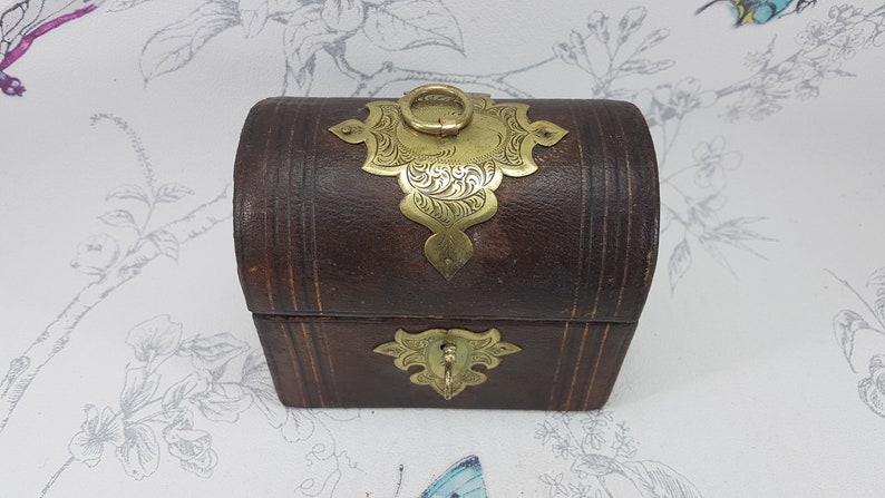 May include: A small, brown leather box with ornate gold metal accents. The box has a hinged lid with a decorative brass handle.