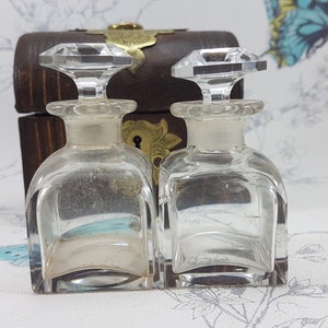May include: Two clear glass bottles with crystal stoppers, sitting on a wooden box with a brass lock and keyhole. The bottles are empty and have a square shape.