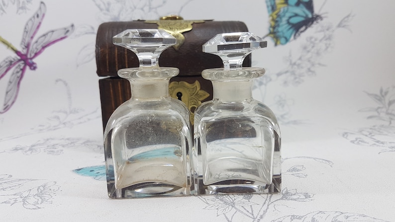 May include: Two clear glass bottles with crystal stoppers, sitting on a wooden box with a brass lock and keyhole. The bottles are empty and have a square shape.