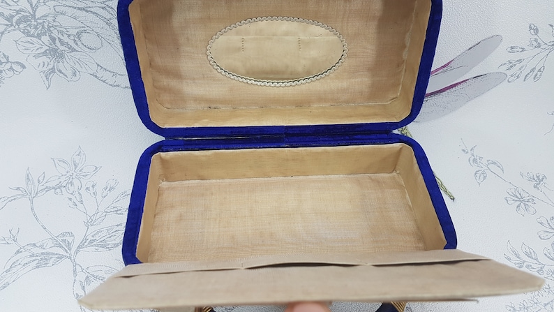 May include: An empty blue velvet jewelry box with a beige interior. The box has a decorative oval design on the lid.