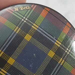 K&ouml;nnte beinhalten: Ein runder Knopf mit einem farbenfrohen Schottenmuster in Blau-, Gr&uuml;n-, Gelb-, Orange- und Lilat&ouml;nen. Der Text "M'Beth" ist in goldfarbenen Buchstaben oben zu sehen. Der Knopf wird von den Fingern einer Person gehalten.