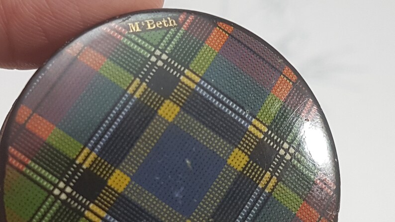 K&ouml;nnte beinhalten: Ein runder Knopf mit einem farbenfrohen Schottenmuster in Blau-, Gr&uuml;n-, Gelb-, Orange- und Lilat&ouml;nen. Der Text "M'Beth" ist in goldfarbenen Buchstaben oben zu sehen. Der Knopf wird von den Fingern einer Person gehalten.