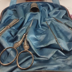 May include: A vintage sewing kit containing silver-toned tools, including scissors, a needle threader, and a thimble, presented on a blue satin background.