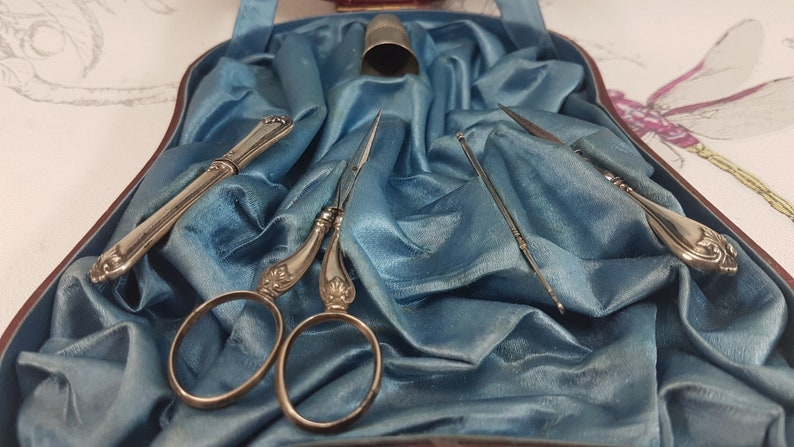 May include: A vintage sewing kit containing silver-toned tools, including scissors, a needle threader, and a thimble, presented on a blue satin background.