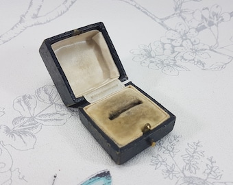 Könnte beinhalten: Eine kleine, schwarze, quadratische Ringbox mit einem Samt-Innenfutter. Die Box ist geöffnet und der Samt ist leicht abgenutzt.