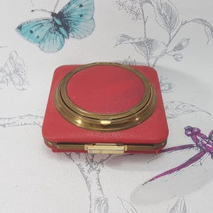 May include: A vintage red compact with a gold-colored rim and clasp. The compact is square-shaped and has a circular top. The background features a butterfly and floral design.