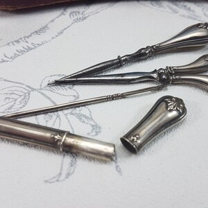 May include: A set of silver sewing tools, including a thimble, a pair of scissors, and two needle threaders. The tools are laid out on a white surface with a floral pattern.