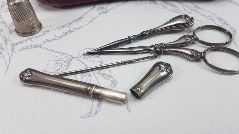 May include: A set of silver sewing tools, including a thimble, a pair of scissors, and two needle threaders. The tools are laid out on a white surface with a floral pattern.