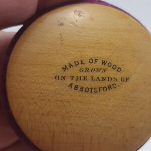 May include: Close-up of a round, wooden object with a purple velvet border. The object has the text "Made of Wood Grown on the Lands of Abbotsford." inscribed on it.