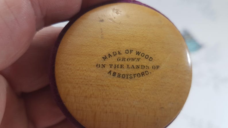 May include: Close-up of a round, wooden object with a purple velvet border. The object has the text "Made of Wood Grown on the Lands of Abbotsford." inscribed on it.