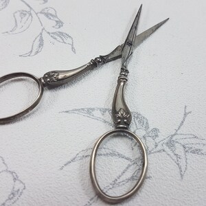 May include: A pair of antique silver scissors with ornate handles and oval-shaped loops. The scissors are closed and lying on a white surface with a floral pattern.