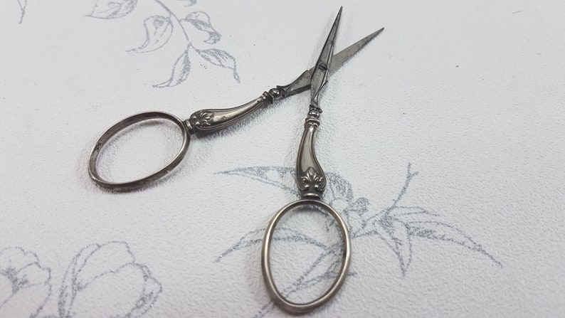 May include: A pair of antique silver scissors with ornate handles and oval-shaped loops. The scissors are closed and lying on a white surface with a floral pattern.