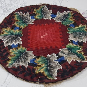 May include: A round, hand-stitched needlepoint cushion with a red centre and a border of green, gold, and white leaves. The cushion is made of wool and is backed with burlap.