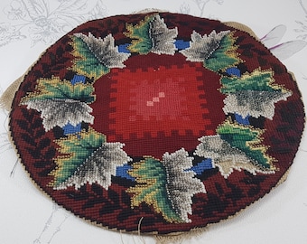 Op de afbeelding: Een rond, met de hand gestikt naaldkunst kussen met een rood midden en een rand van groene, gouden en witte bladeren. Het kussen is gemaakt van wol en is gevoerd met jute.
